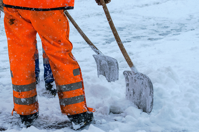 Lower half of person wearing fluorescent orange work trousers and holder shovel in snow
