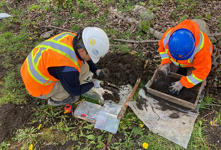 Two people with hard hats and hi-vis jackets kneel over a small dig site and inspecting the earth