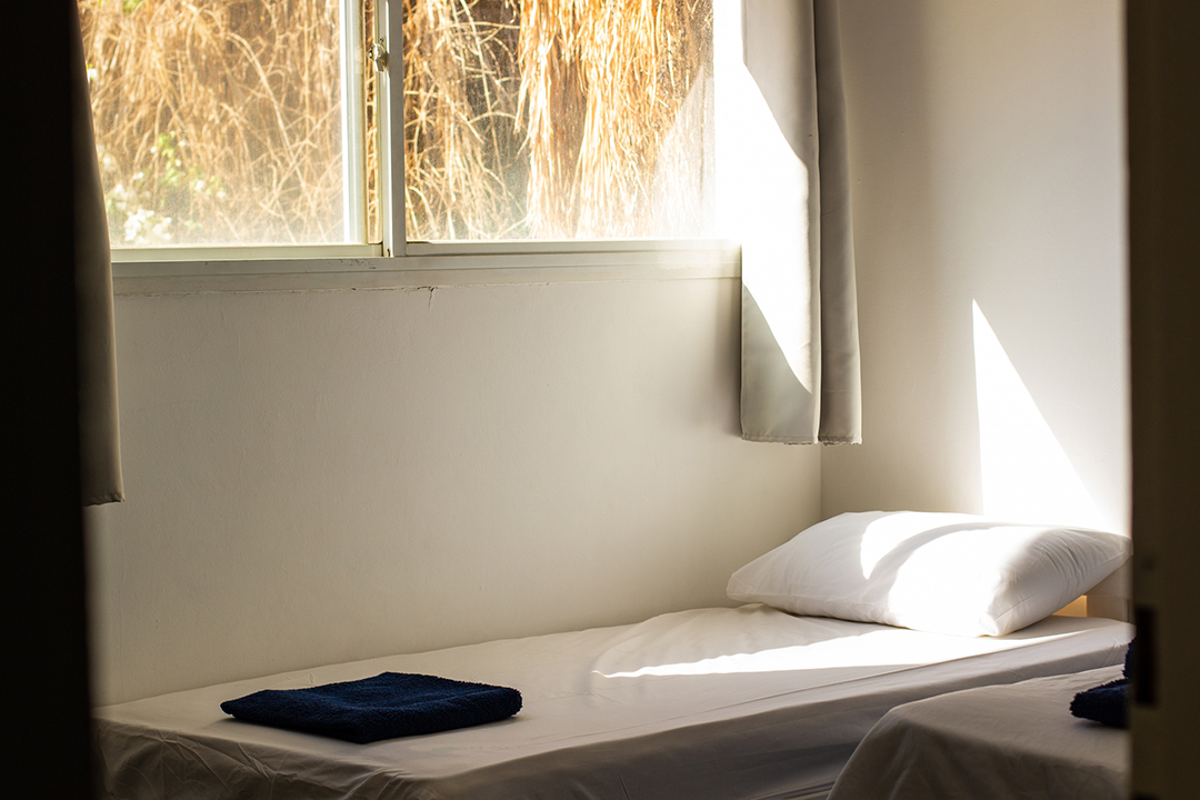 Single bed with pillow and folded towel in a temporary shelter room, illuminated by natural light from a nearby window.
